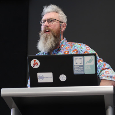 bearded man speaking, wearing a colourful shirt, with laptop and lectern in foreground